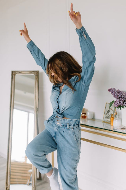 A woman in a denim jumpsuit joyfully dancing with her arms raised in a bright, modern room