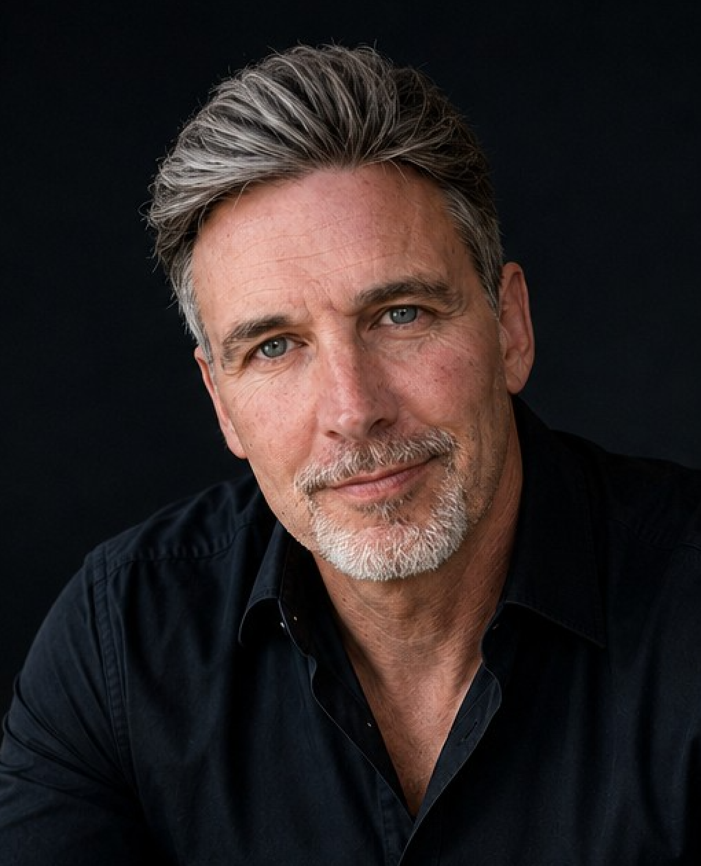 A close-up portrait of Neil Sunley with gray hair and a salt-and-pepper beard, wearing a black button-down shirt against a dark background.